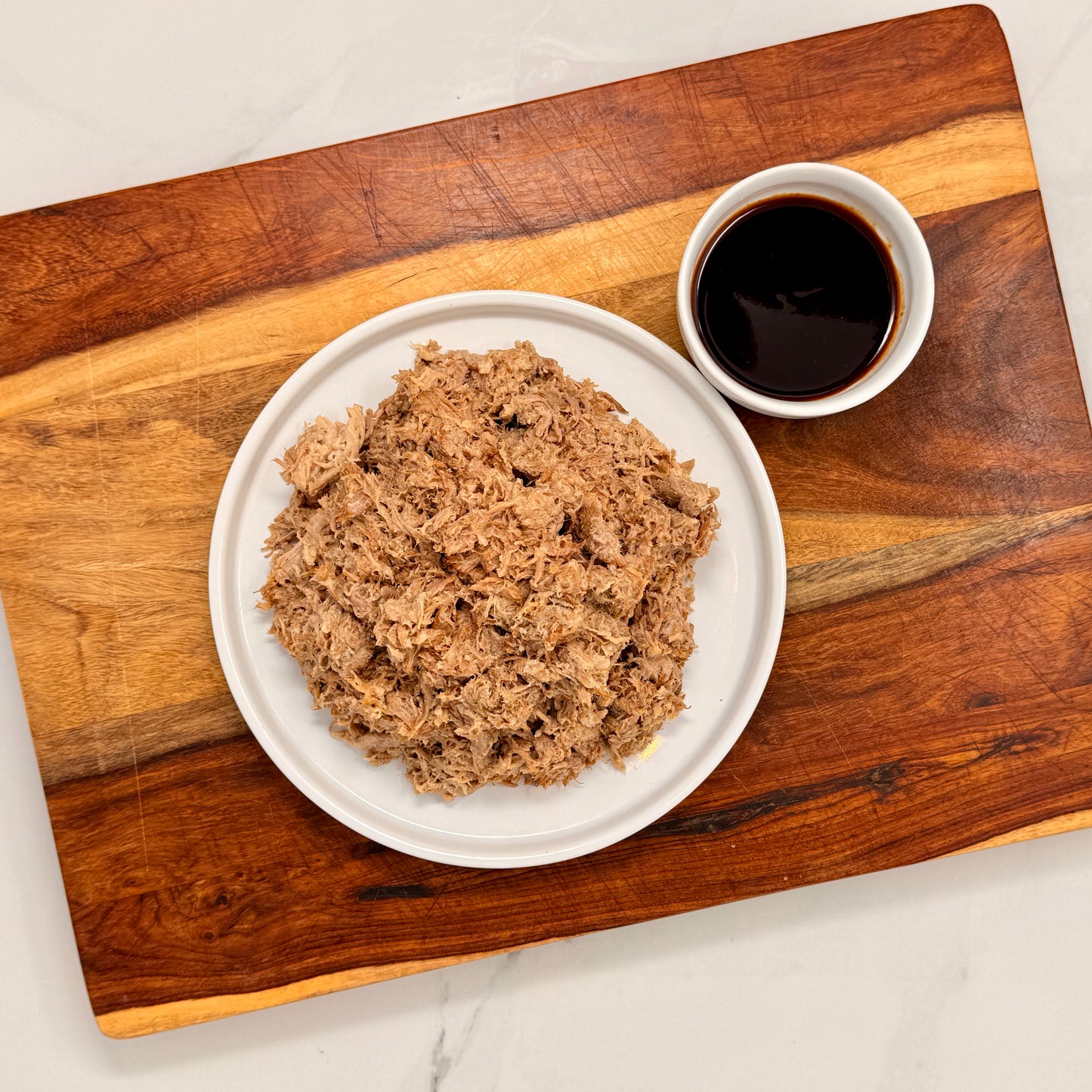 Plated dish of pulled pork on a wooden cutting board with a small bowl of sauce.