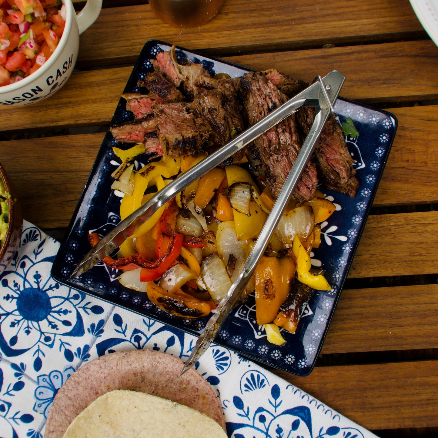 Plated flank steak fajita with tongs, avocado, and pico de gallo on a wooden table.