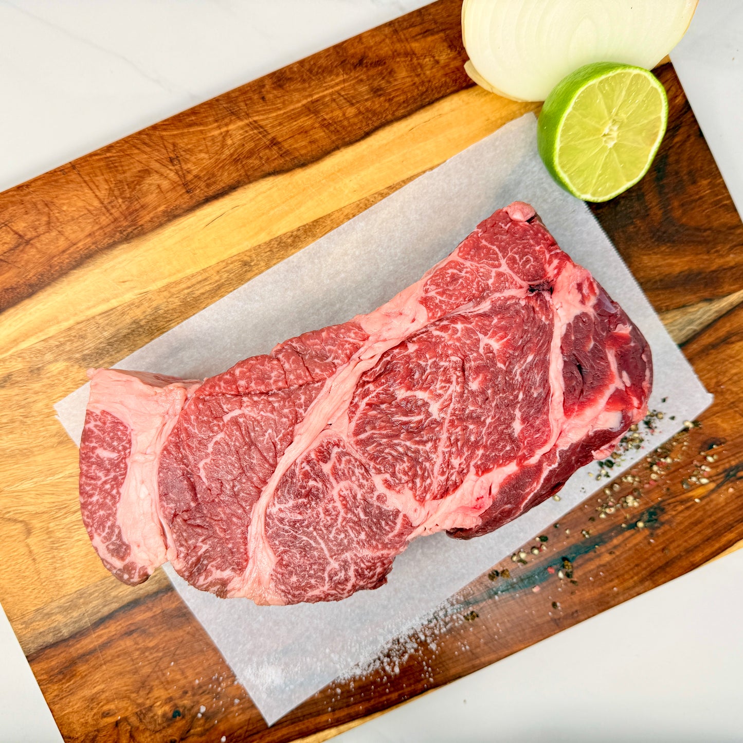 An American Wagyu Chuck Roast from Verlinde Farms on a wooden cutting board with white paper underneath, a yellow onion and lime, as well as salt and pepper are garnishing the board.