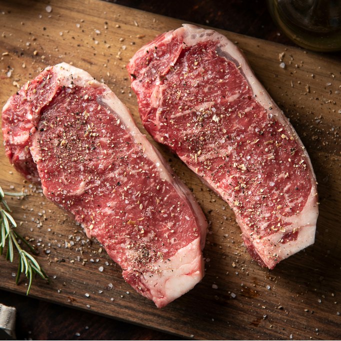 Two raw new york strip steaks on a wooden cutting board with seasonings and herbs.
