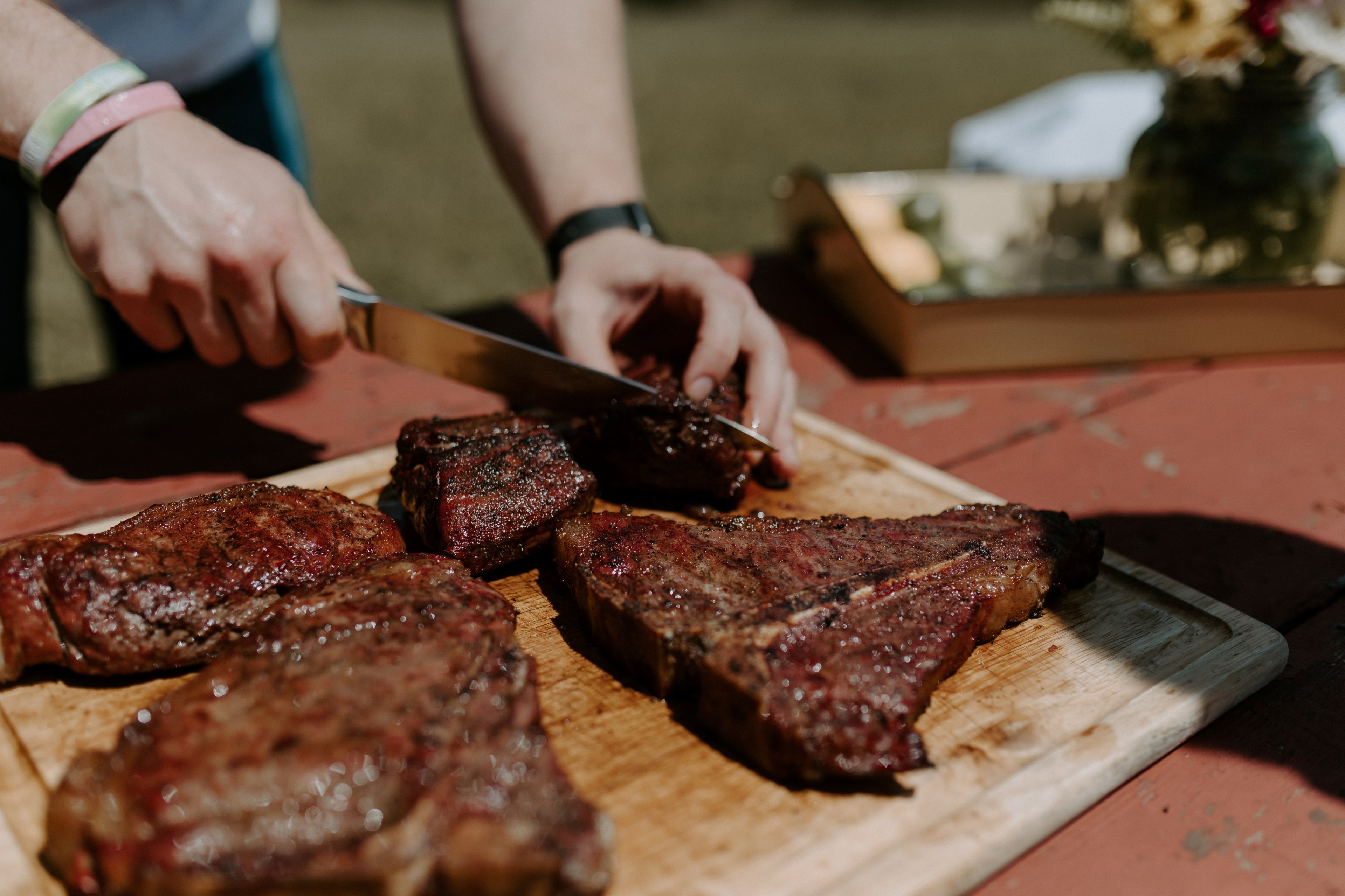 Four perfectly cooked steaks arranged on a cutting board. The cutting board is on a picnic table outside and someone is cutting the steaks with a chef's knife.