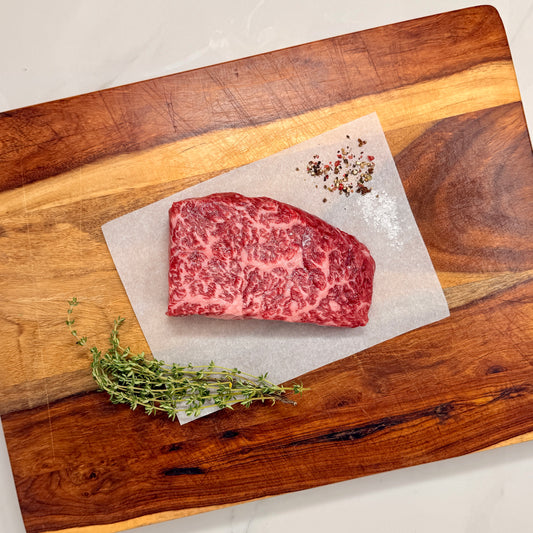 A raw American Wagyu Denver Steak on a wooden cutting board with herbs and seasonings.