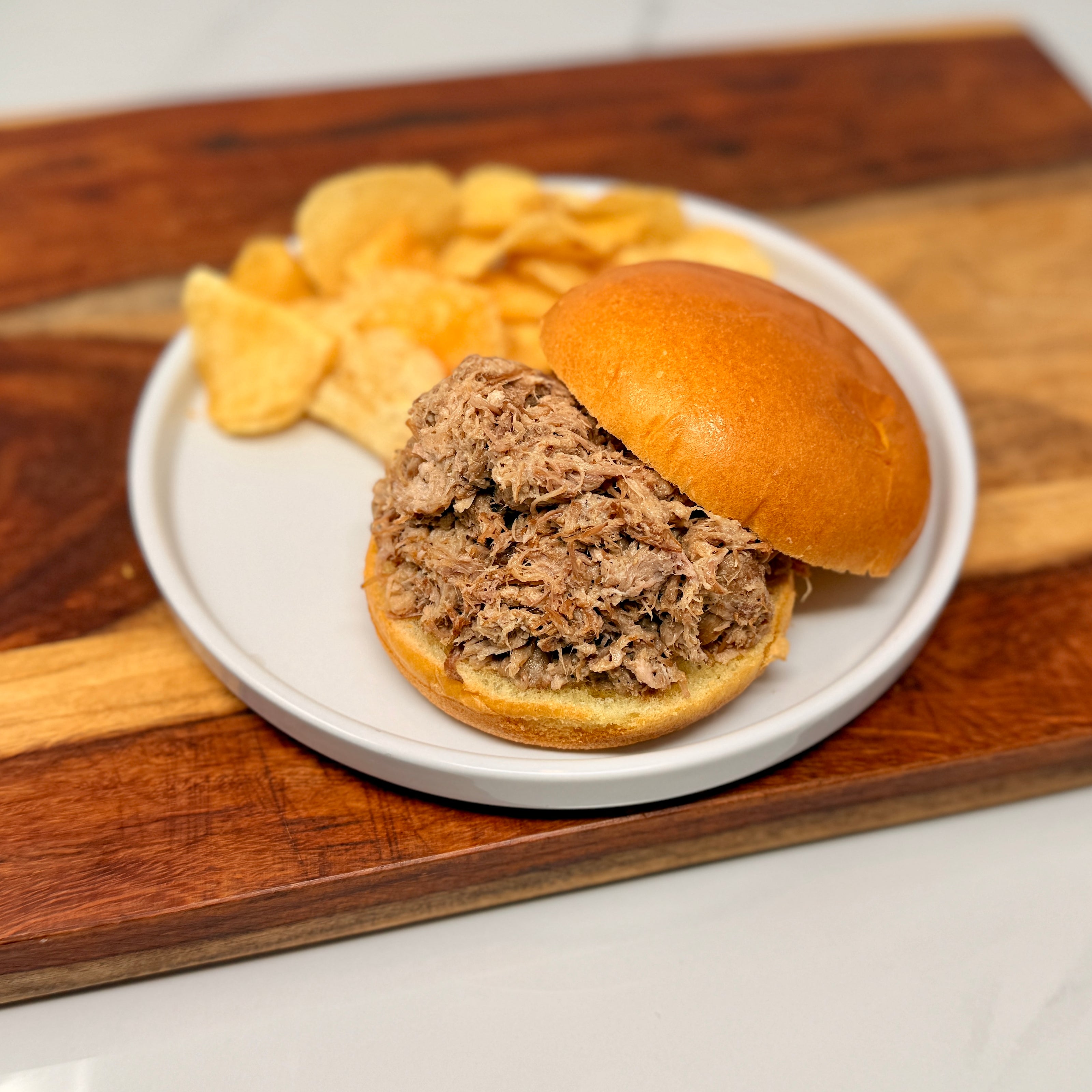 Pulled pork sandwich on a bun with chips on a white plate, placed on a wooden cutting board.