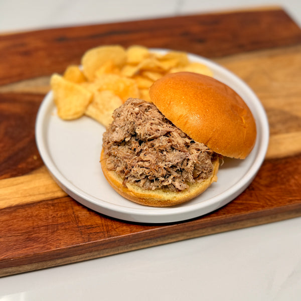Pulled pork sandwich on a bun with chips on a white plate, placed on a wooden cutting board.