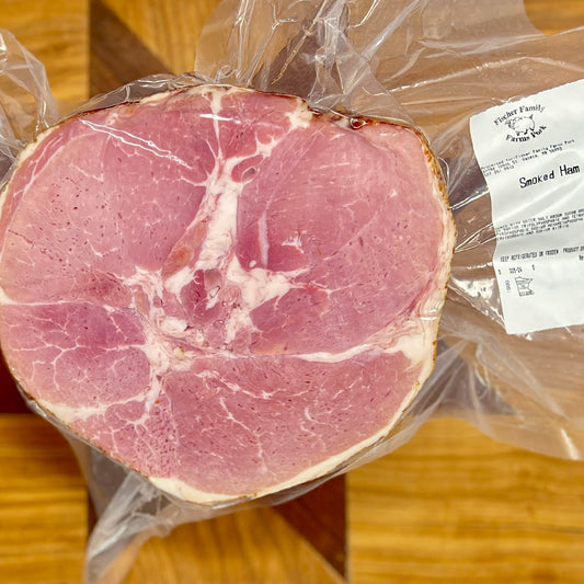 A smoked ham showing off the marbling of heritage breed pork from Fischer Family Farms in clear plastic wrap on a wooden surface