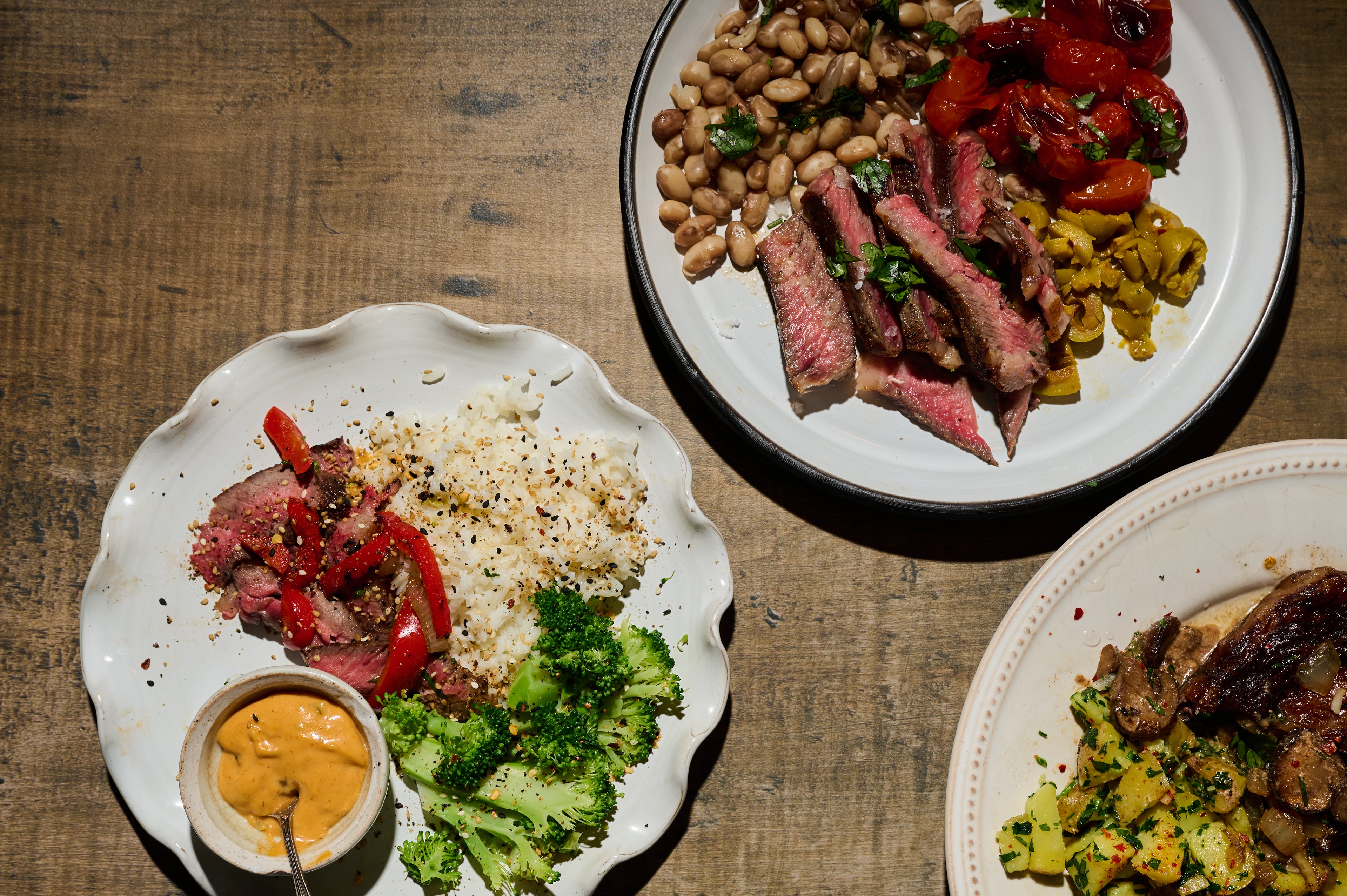 Two plates of sliced New York Strip Steaks, cooked medium rare, and served with vegetables, and sides on a wooden table.