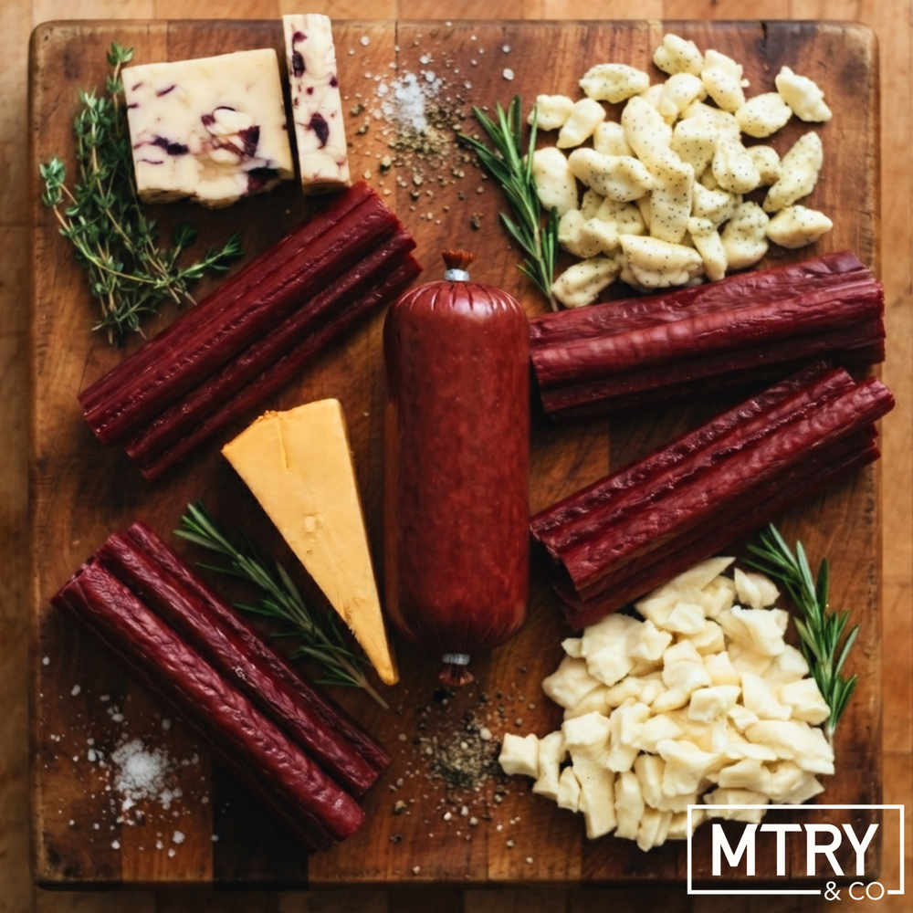 A styled charcuterie board featuring a whole summer sausage, beef sticks, a wedge of gouda cheese, cheese curds, and a block of cranberry cheddar cheese, garnished with rosemary and salt on a wooden serving board. The 'MTRY & CO' logo is visible in the bottom right corner.