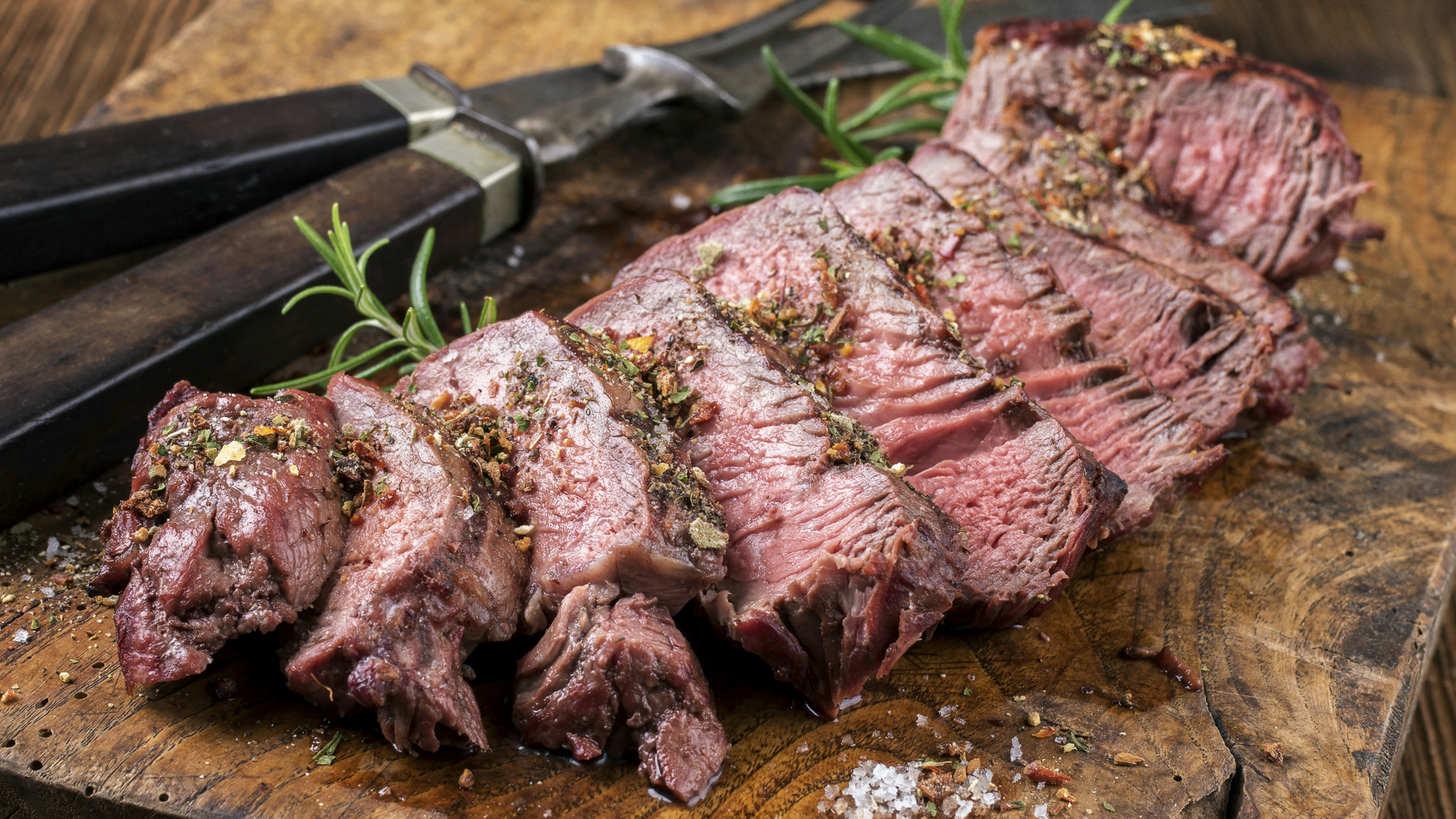 Sliced steak on a wooden cutting board with herbs and a knife.