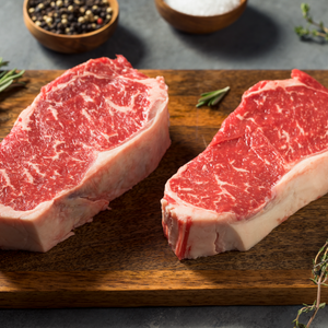 Two raw steaks on a wooden cutting board with spices in the background.