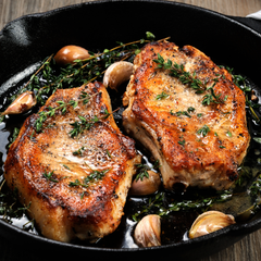 two pan seared pork chops in a cast iron grill with garlic and thyme