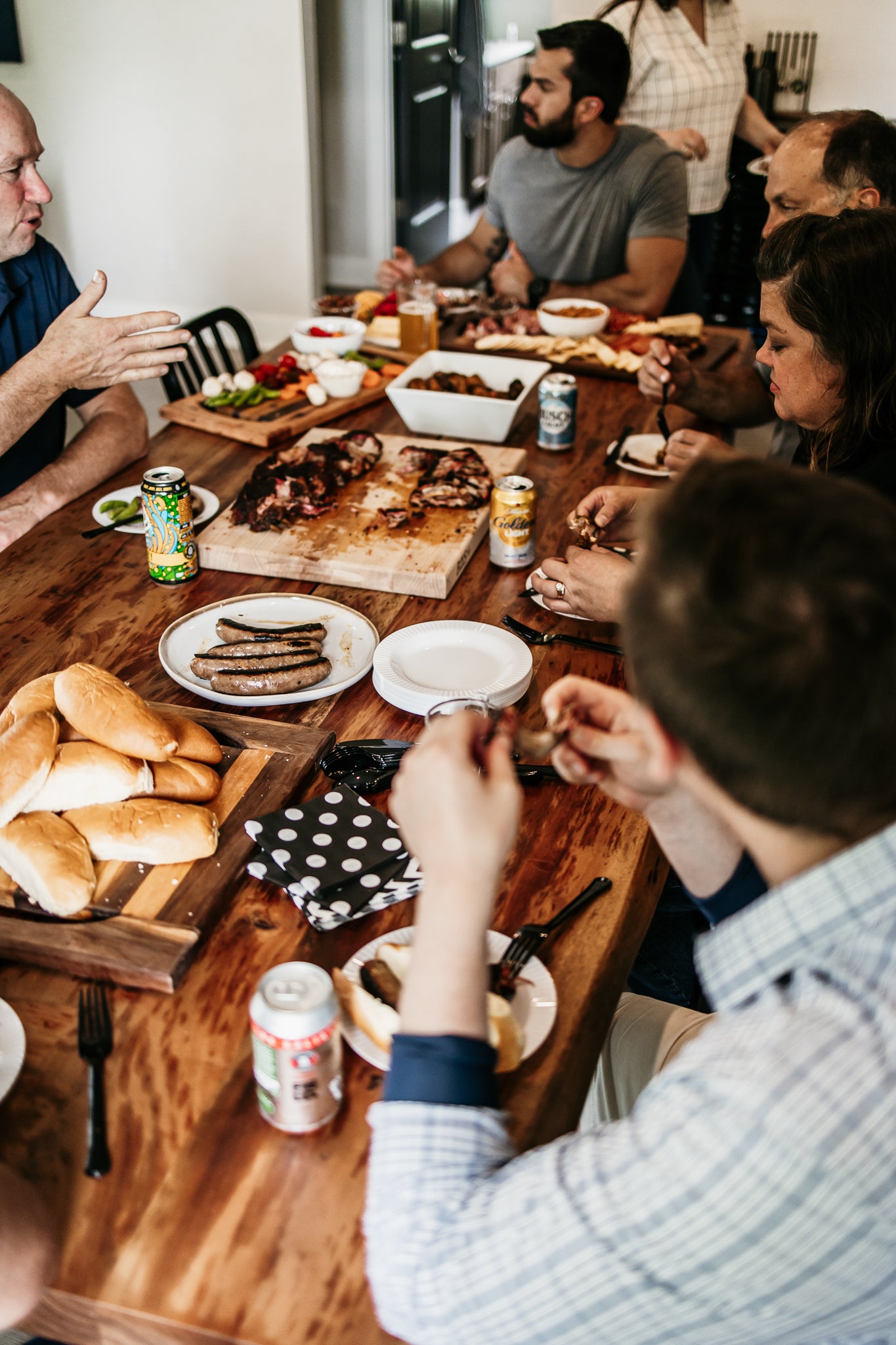 MTRY & CO Crew and Friends gathered around a wooden dining table for a meal. Bratwurst, steaks, chicken wings, and more are on the table.