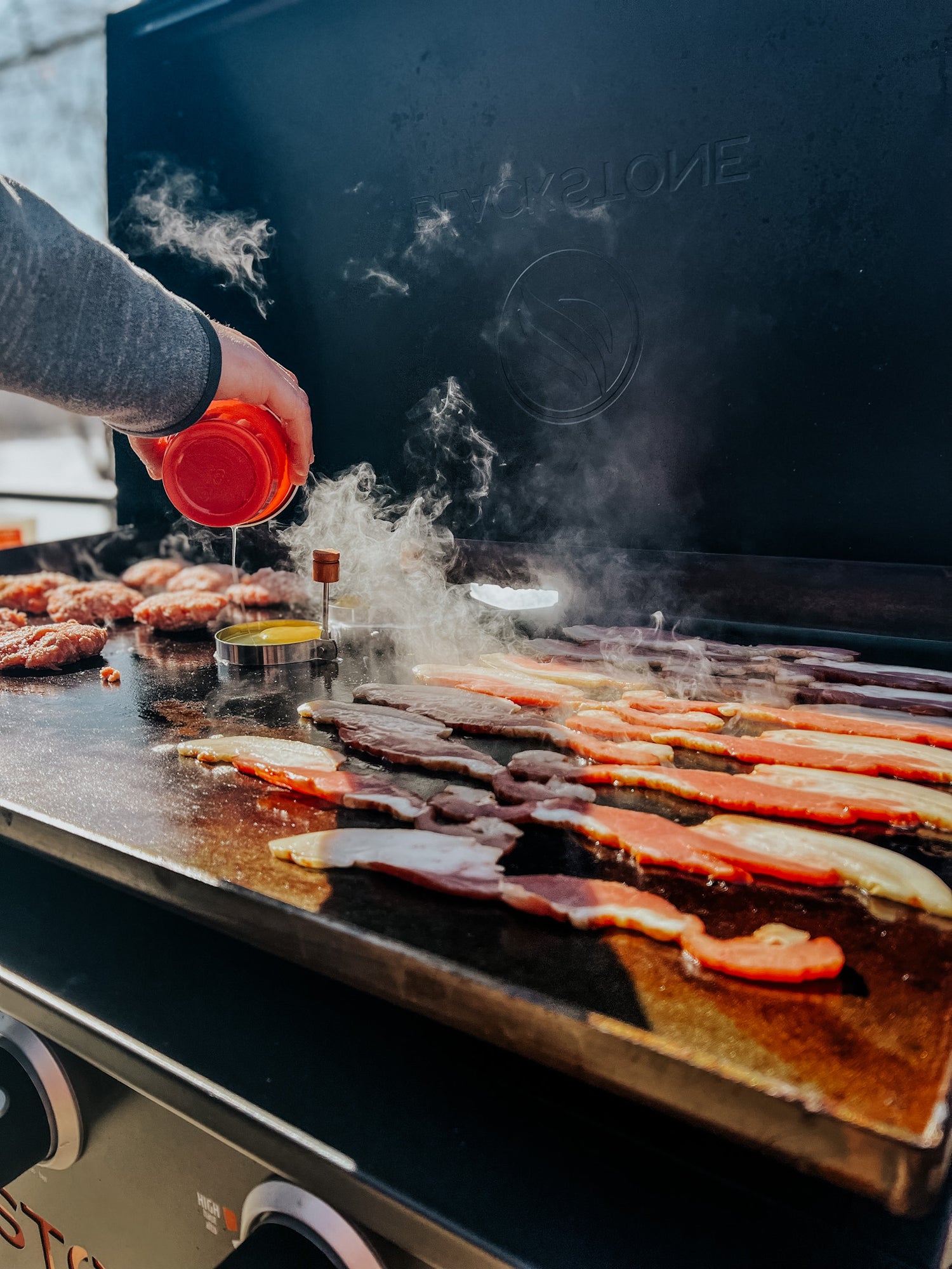 Person grilling outdoors by a lake, there are strips of bacon, eggs in ring molds, breakfast sausage, and patties being fried. Steam is pouring off the grill in the cold air.