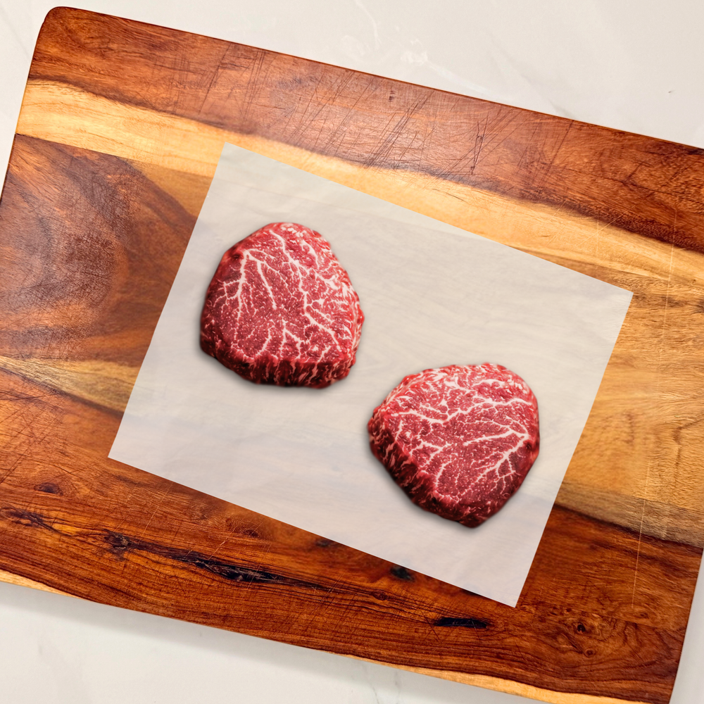 Two raw, well-marbled wagyu beef tenderloin filets on a sheet of parchment paper, placed on a wooden cutting board.