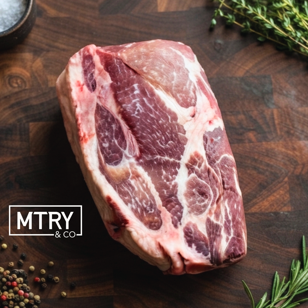 A heritage pork roast from Sixth Day Farms highlighting the rich, marbled meat of heritage breed pork. The roast is on a dark wood cutting board with a garnish of fresh herbs and peppercorns.