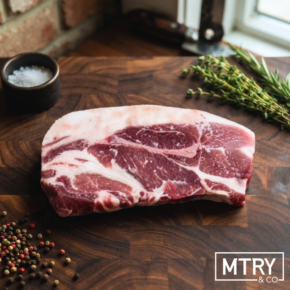 A heritage pork steak from Sixth Day Farms sitting on a dark wood cutting board. The steak has a thick cap of rich heritage pork fat and shows of the marbling characteristic of heritage breed pork. It is surrounded by fresh herbs and peppercorns.