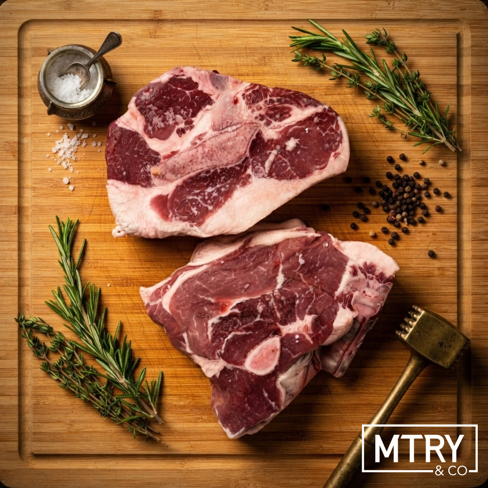 A heritage pork steak from Sixth Day Farms sitting on a dark wood cutting board. The steak has a thick cap of rich heritage pork fat and shows of the marbling characteristic of heritage breed pork. It is surrounded by fresh herbs and peppercorns.