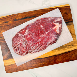 A raw, marbled, American Wagyu Flank Steak on a sheet of white parchment paper atop a wooden cutting board on a white marble counter.