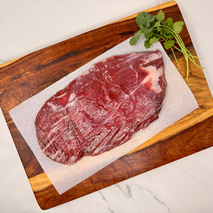 A raw, marbled, American Wagyu Flank Steak with a few sprigs of cilantro on a sheet of white parchment paper atop a wooden cutting board on a white marble counter.