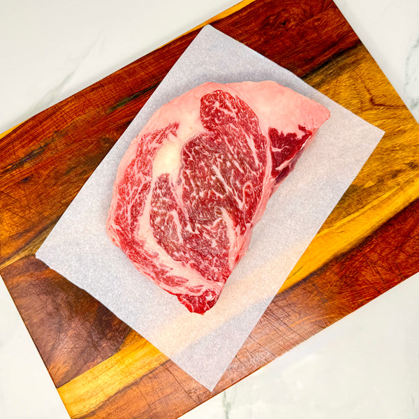 A raw American Wagyu ribeye steak on a sheet of white parchment paper, placed on a wooden cutting board over a white marble countertop.