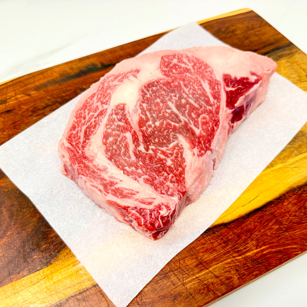 A raw American Wagyu ribeye steak on a sheet of white parchment paper, placed on a wooden cutting board over a white marble countertop.