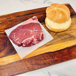 An American Wagyu sirloin steak, raw, on parchment paper next to a sliced kaiser roll on a wooden cutting board, set on a white marble countertop.