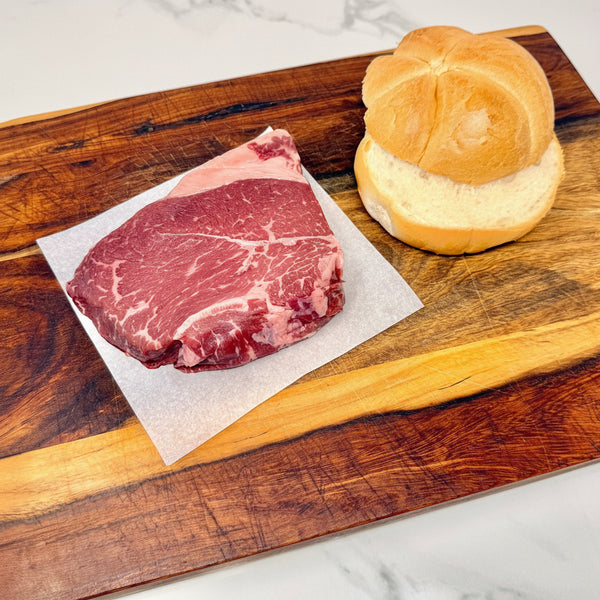 An American Wagyu sirloin steak, raw, on parchment paper next to a sliced kaiser roll on a wooden cutting board, set on a white marble countertop.