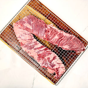 A single, large, American Wagyu skirt steak with visible marbling, placed on a metal wire cooling rack over a cutting board on a white marble surface.