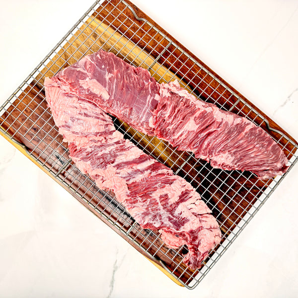 A single, large, American Wagyu skirt steak with visible marbling, placed on a metal wire cooling rack over a cutting board on a white marble surface.