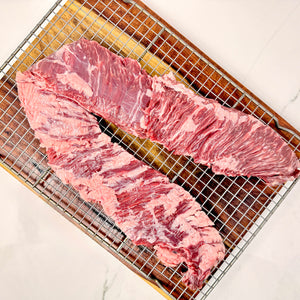 A single, large, American Wagyu skirt steak with visible marbling, placed on a metal wire cooling rack over a cutting board on a white marble surface.