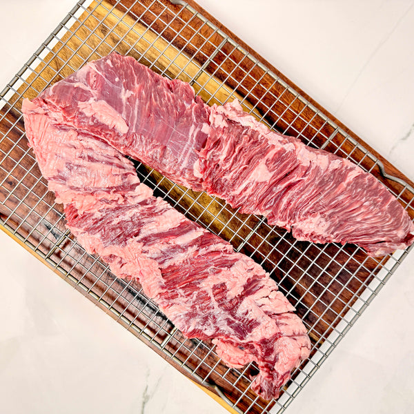 A single, large, American Wagyu skirt steak with visible marbling, placed on a metal wire cooling rack over a cutting board on a white marble surface.