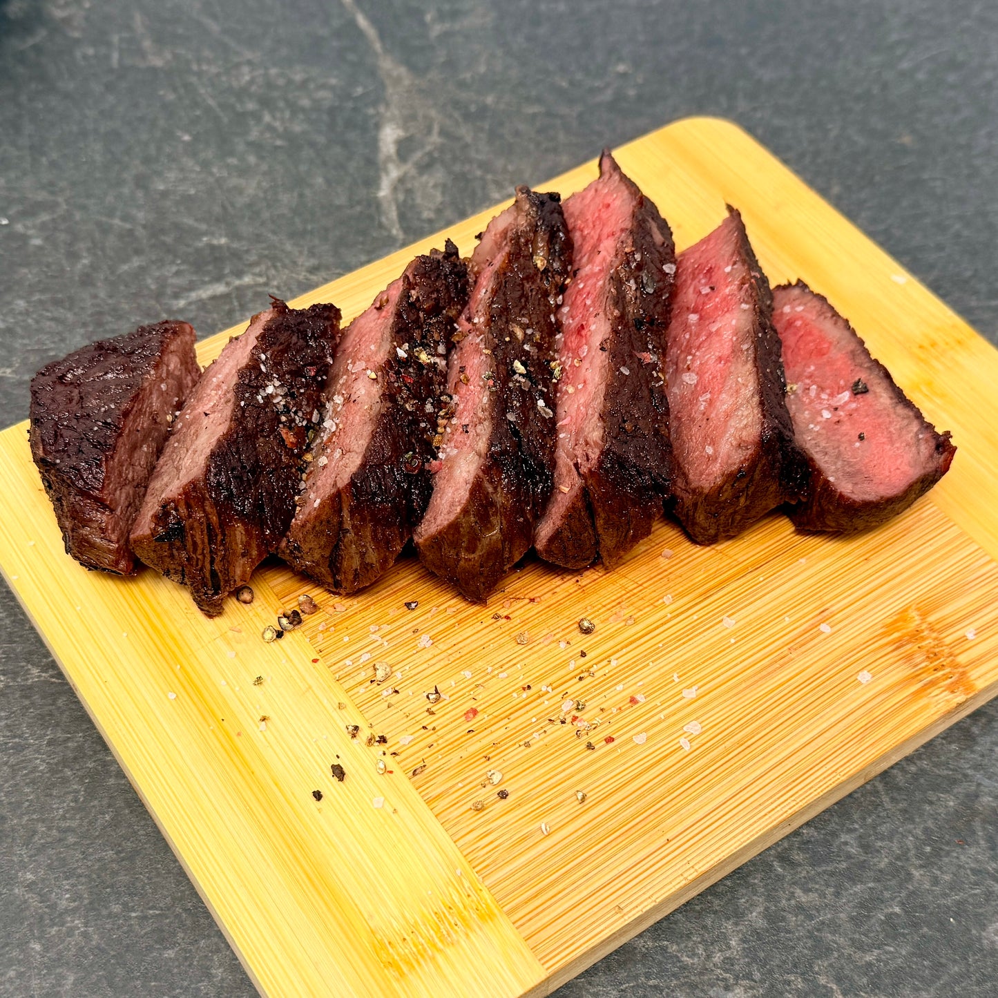 Sliced cooked American Wagyu Denver Steak on a wooden cutting board with a dark background