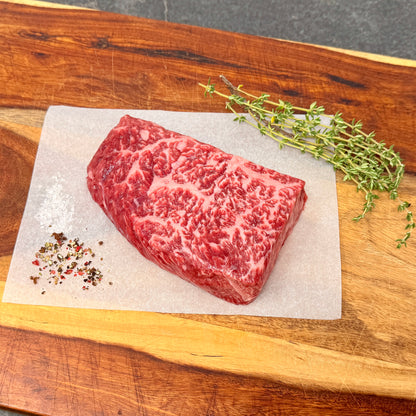 A raw American Wagyu Denver Steak on a wooden cutting board with thyme, salt, and pepper.