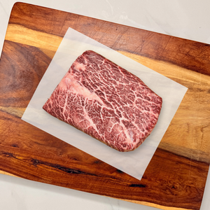 A well-marbled American Wagyu flat iron steak resting on a sheet of white parchment paper atop a wooden cutting board on a white marble countertop.