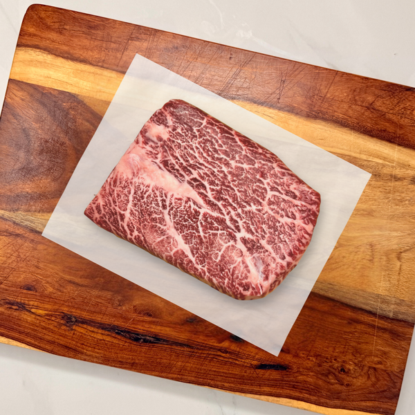A well-marbled American Wagyu flat iron steak resting on a sheet of white parchment paper atop a wooden cutting board on a white marble countertop.